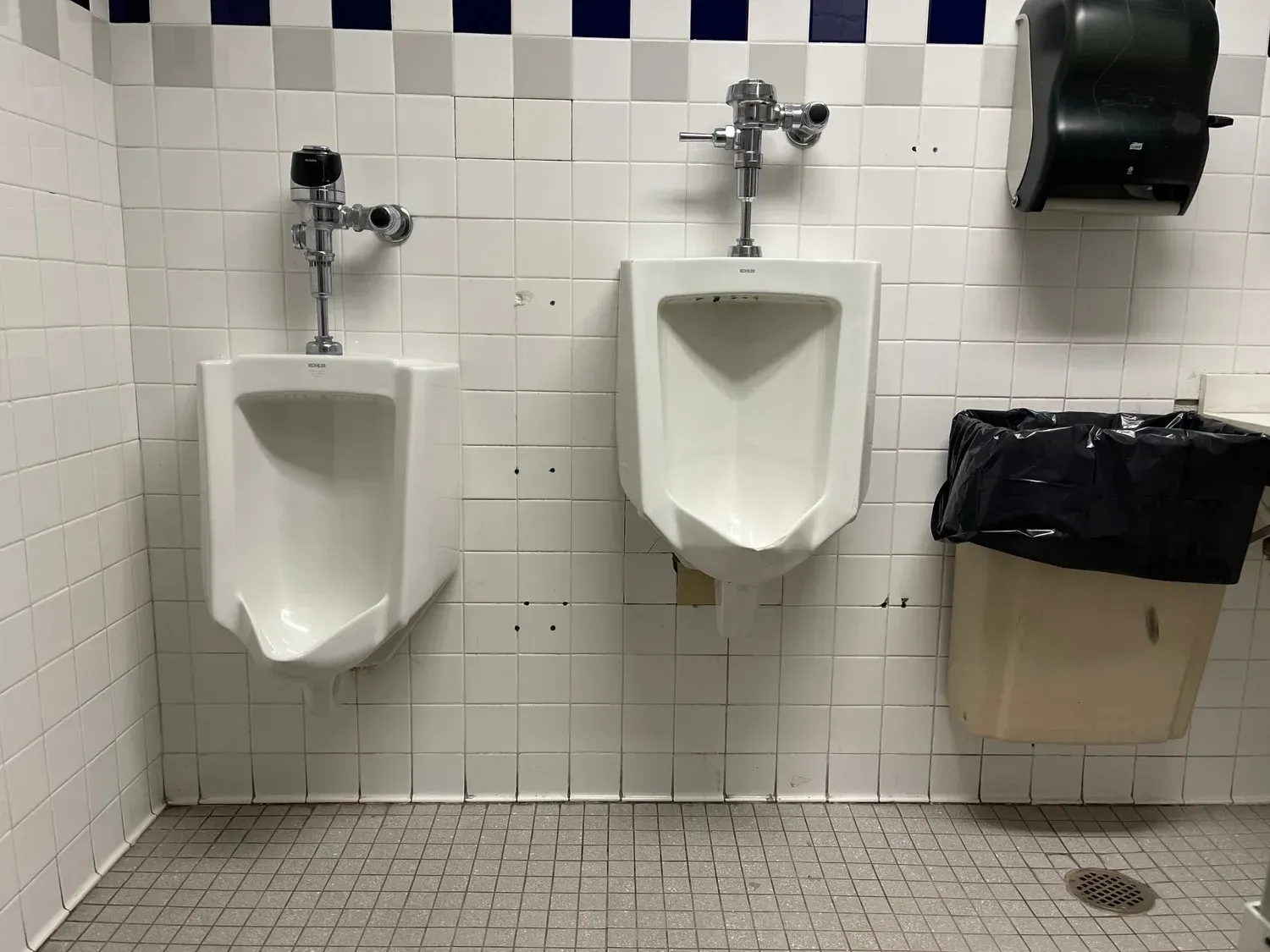 Public restroom with two white urinals, tiled walls, and a black paper towel dispenser.
