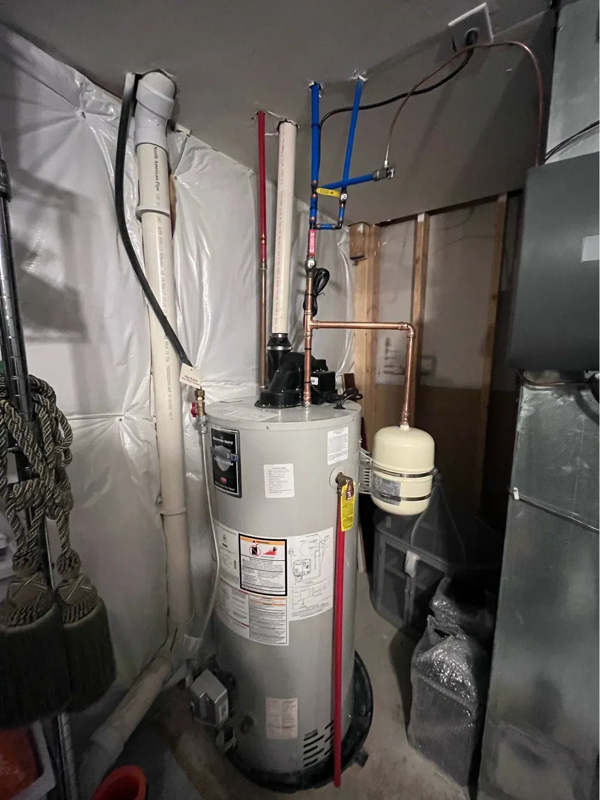 Water heater and plumbing in a cramped utility closet with exposed pipes and insulation.