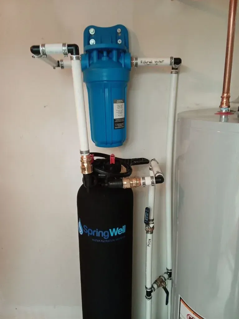 Blue water filter mounted on black SpringWell tank beside copper pipes and a white water heater.