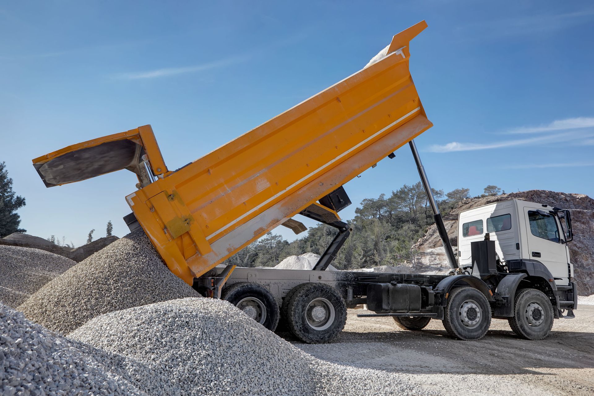 Yellow dump truck unloading gravel at a quarry under a blue sky.