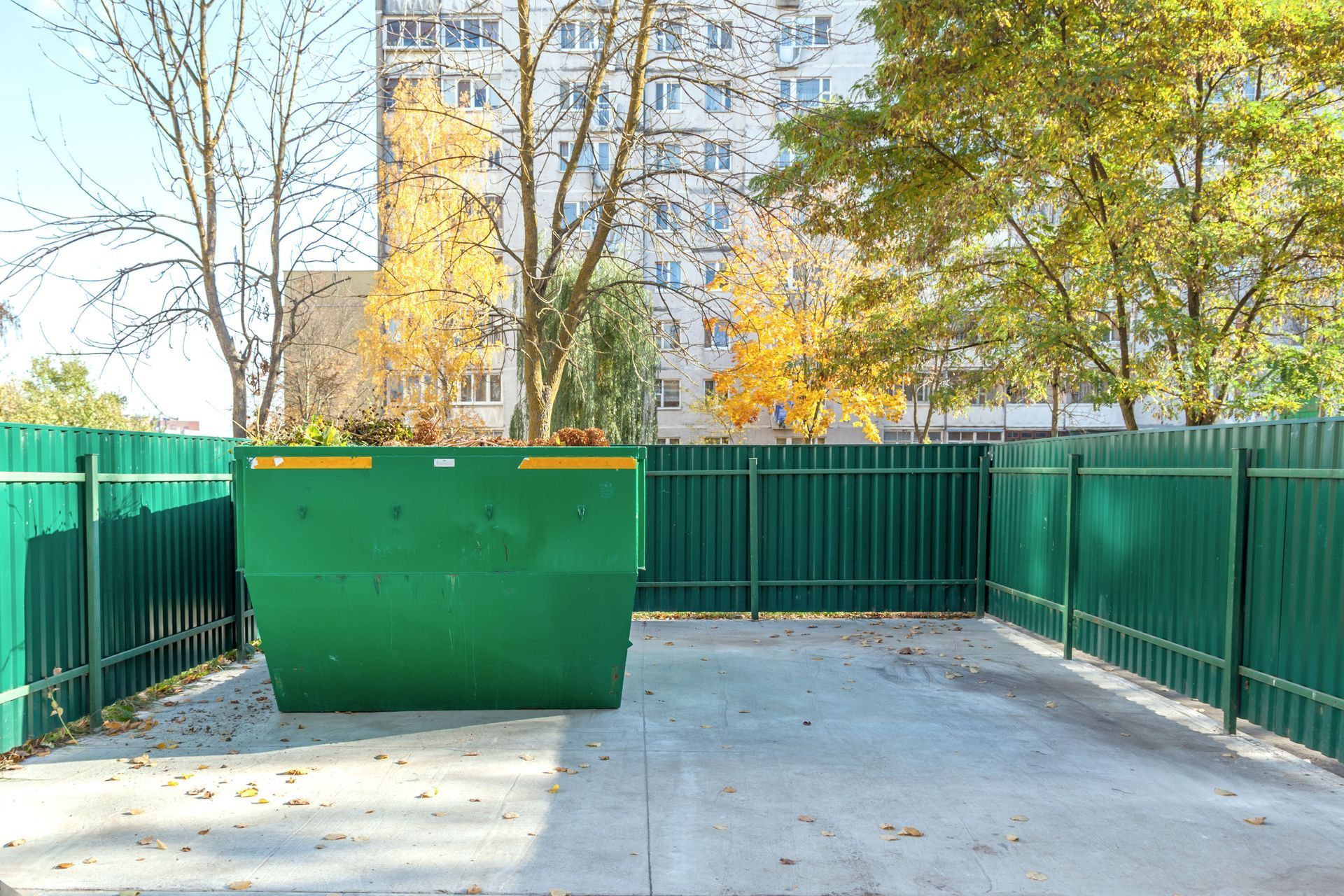 Green dumpster in a fenced area, concrete ground. A building and trees are in the background.