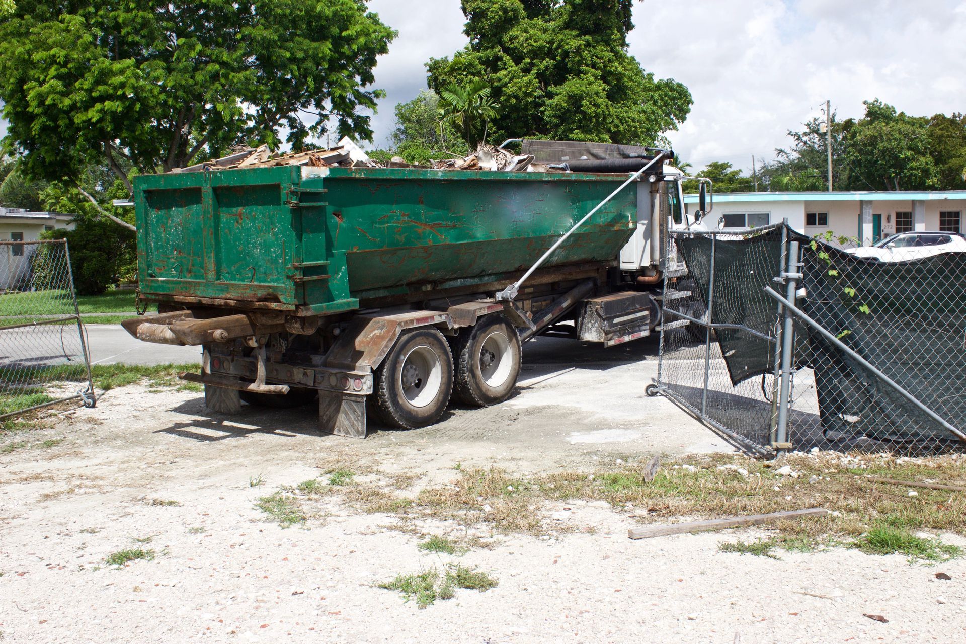 Green dumpster on a truck, filled with debris, parked on gravel in front of a chain link fence.