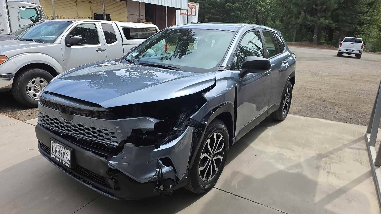 Dark gray damaged SUV parked in a driveway, front-left view, with another truck and a gravel lot in the background