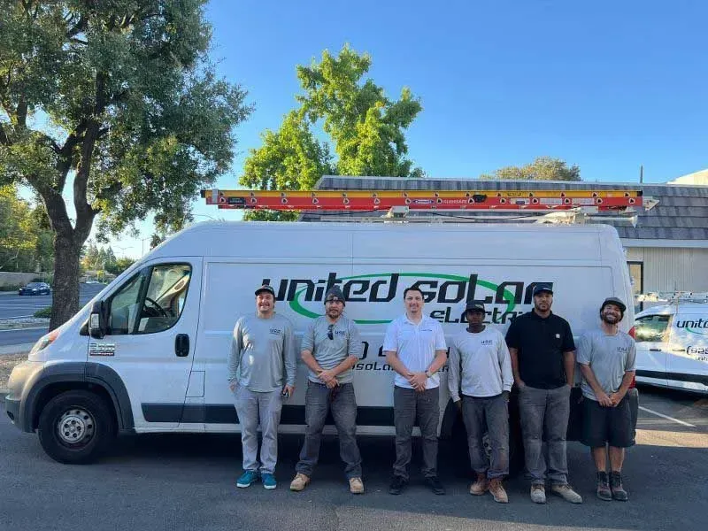 Six men standing in front of a white service van with ladders on top, outdoors in daylight