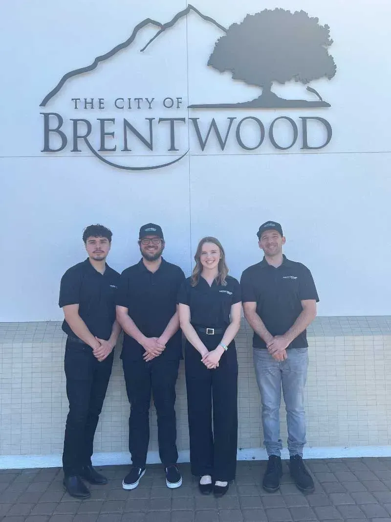 Four people stand in front of The City of Brentwood sign, wearing dark clothing on a paved walkway.