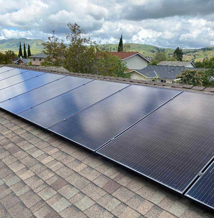 Solar panels mounted on a shingled rooftop under cloudy skies, with houses and hills in the background