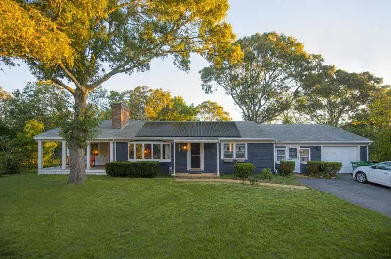 Single-story blue-gray house with a large front lawn, mature trees, and a driveway at sunset