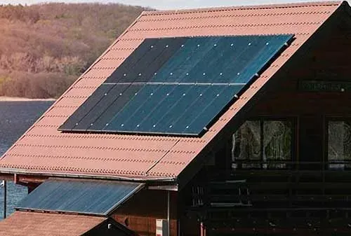 Solar panels mounted on a red-roofed house by a lakeside wooded hillside