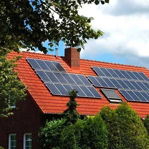 Red-tiled house roof with solar panels under a blue sky, partly framed by trees