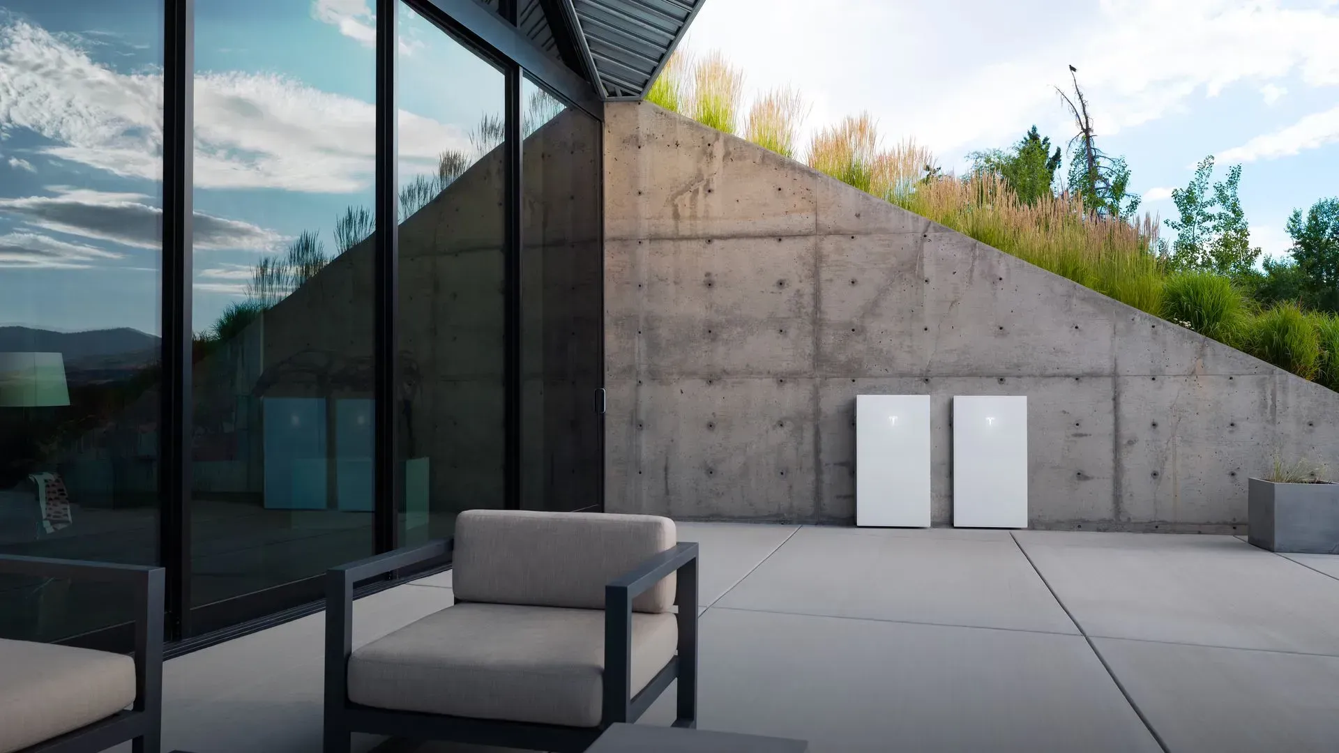 Modern concrete patio with lounge chair, glass wall, and sloped retaining wall against a cloudy sky
