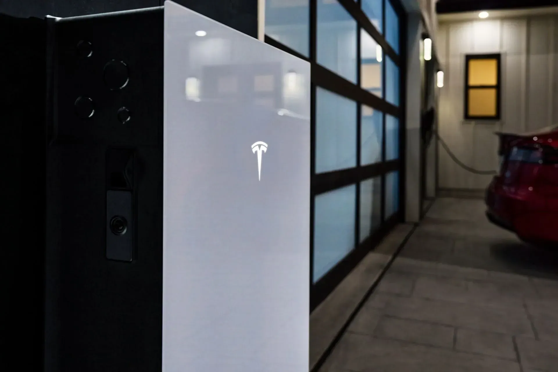 White Tesla charging station beside a garage at dusk, with a red car parked nearby.