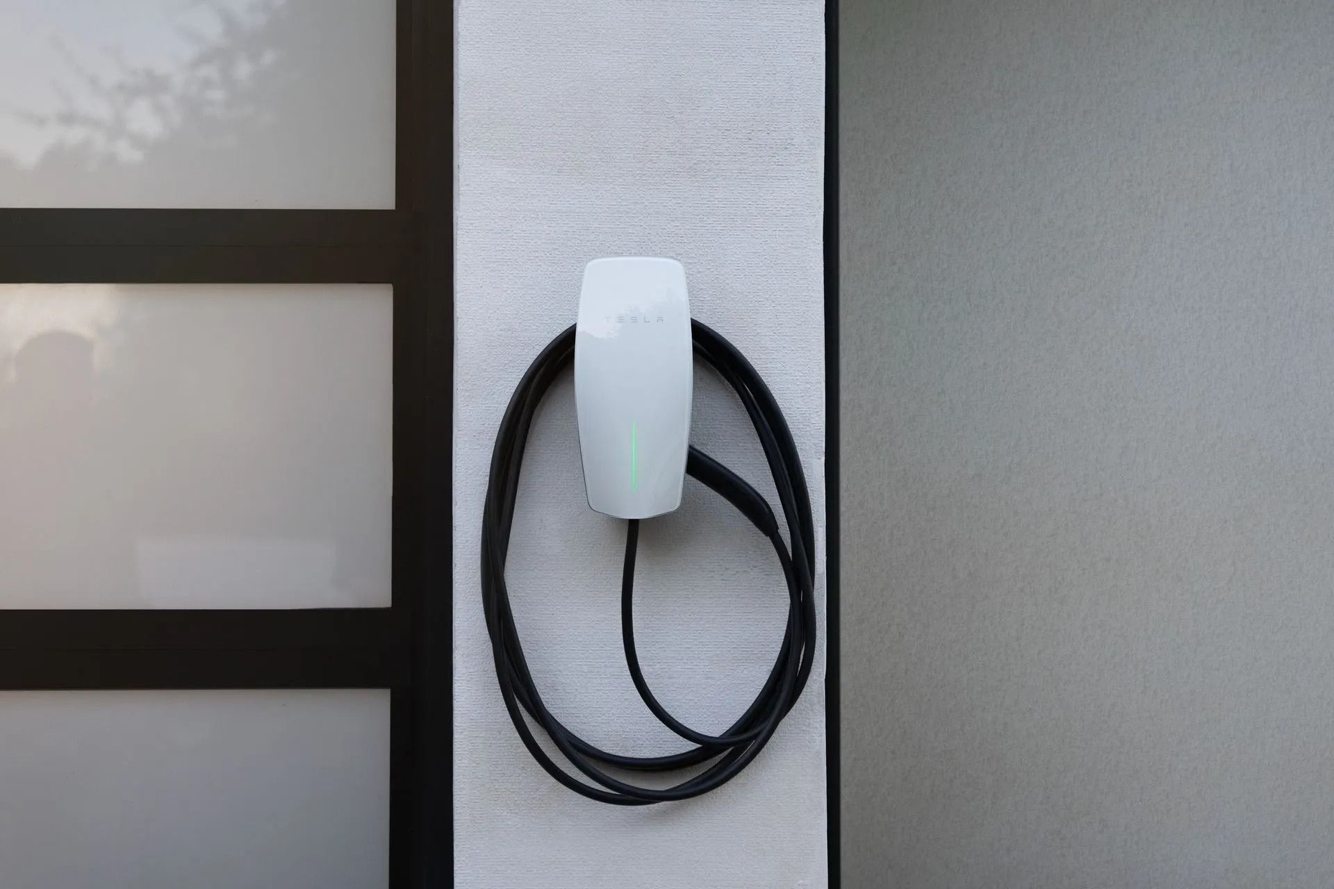 White wall-mounted EV charger with coiled black cable beside a window.