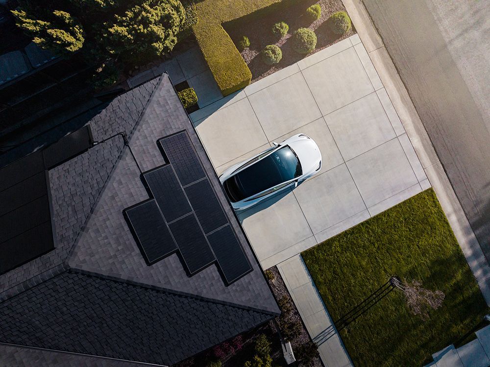 Aerial view of a white car parked in a driveway beside a dark-roofed house and green lawn.