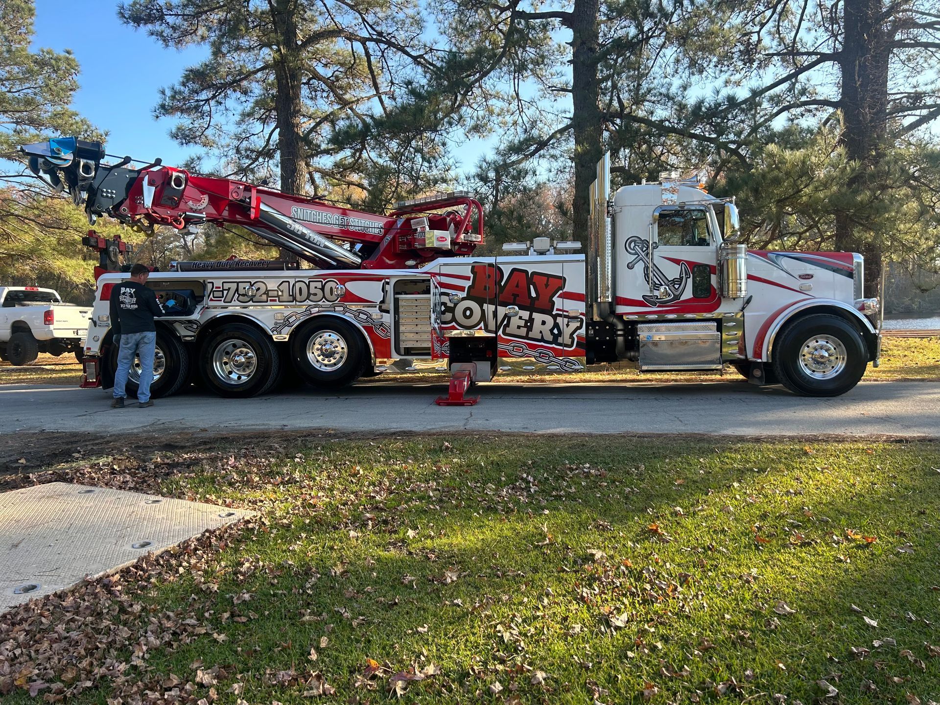A red and white tow truck parked on a paved road with a person standing beside it, set against a backdrop of trees.