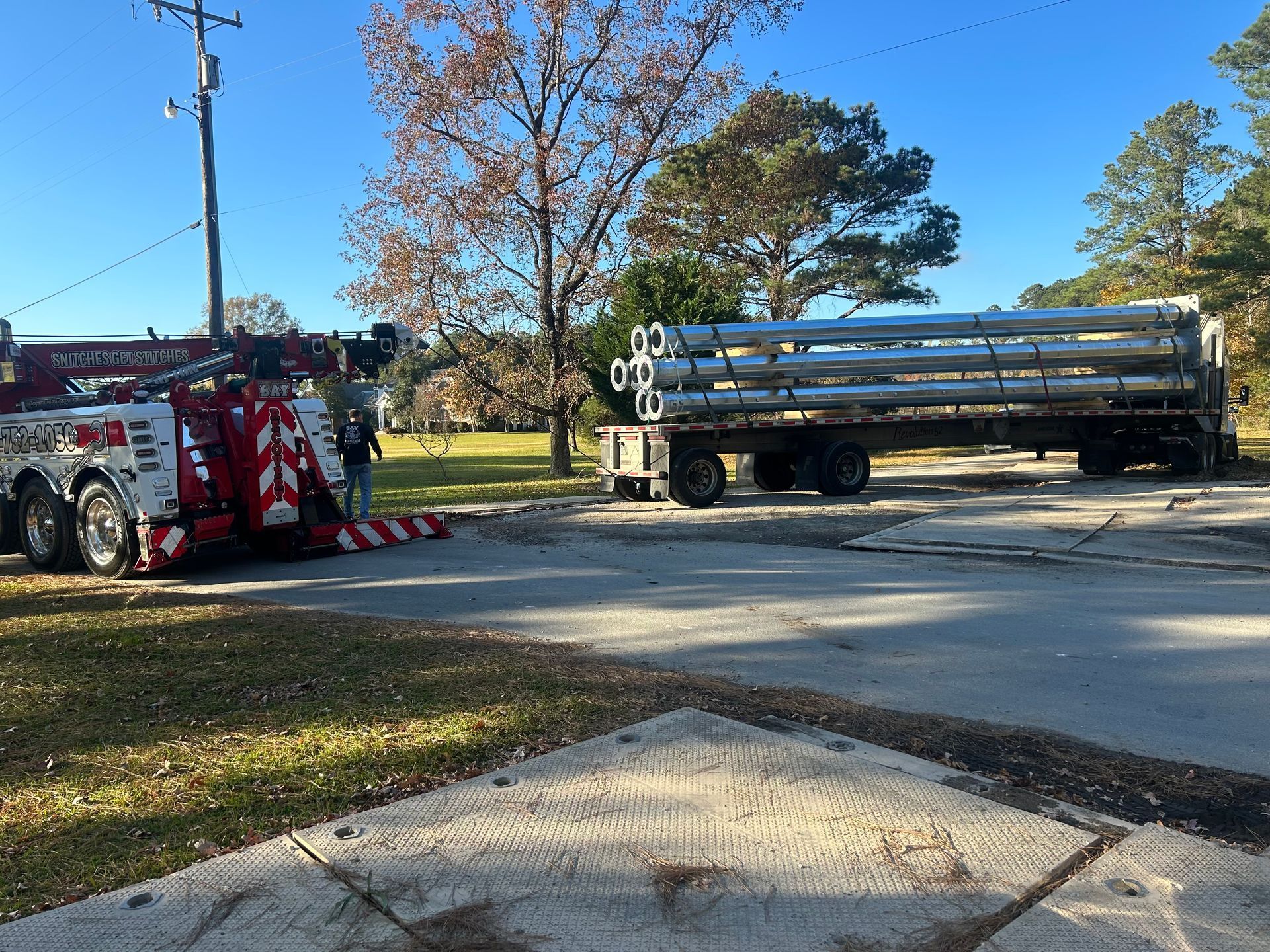 A tow truck parked on a road next to a flatbed trailer carrying a load of metal pipes on a sunny day.