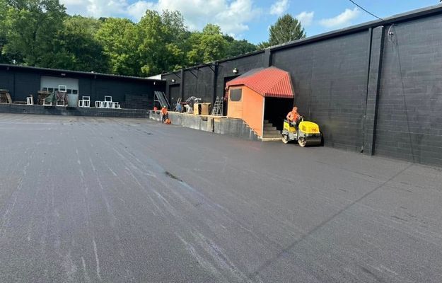 Black asphalt parking lot being paved near a dark industrial building. Workers and machinery are present.