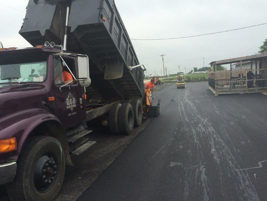 A dump truck unloading asphalt onto a road surface, with workers present; overcast sky.