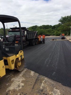 Asphalt paving in progress. Dump truck, road roller, and workers on a black surface.