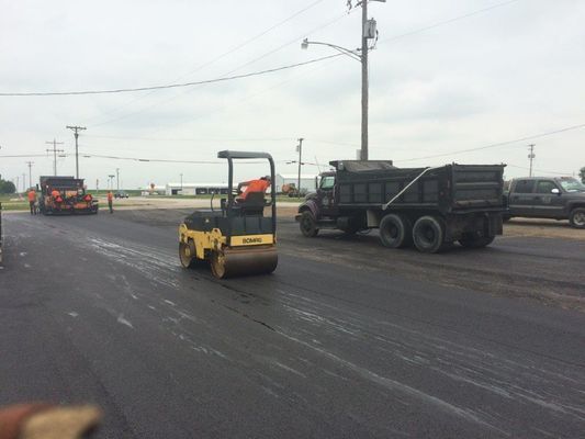 Road paving: small roller, dump truck, paver on asphalt under cloudy sky.