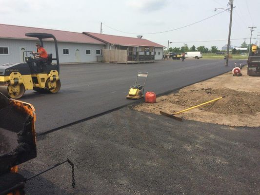 Asphalt paving in progress at a building. A roller, compactor, and workers lay the black surface.