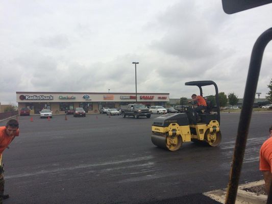 Construction crew paving a parking lot with a yellow roller; overcast sky, businesses in the background.