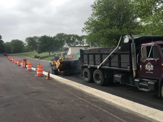 Asphalt paving in progress. A dump truck delivers material to a paving machine on a road. Orange cones line the work area.
