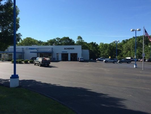 Car dealership with vehicles parked in front of a building. Blue poles and trees are visible against a blue sky.