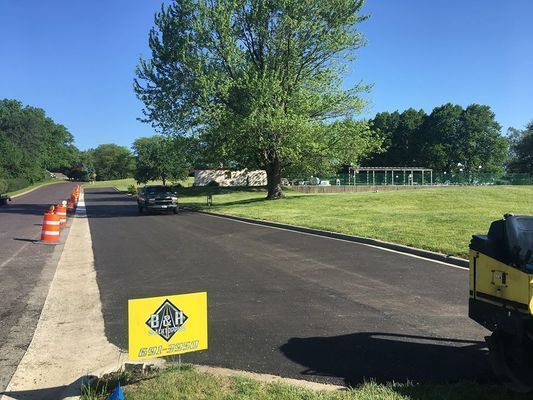 Road resurfacing in progress: black asphalt, orange cones, car, and sign on grassy area with tree.