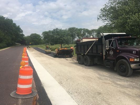 Asphalt road construction: dump truck, paving machine, cones, and freshly paved road.