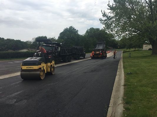 Asphalt paving with rollers, asphalt spreader, and trucks on a road.