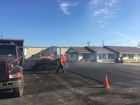 Asphalt paving in progress: worker guiding a machine, truck, buildings, blue sky, traffic cone.