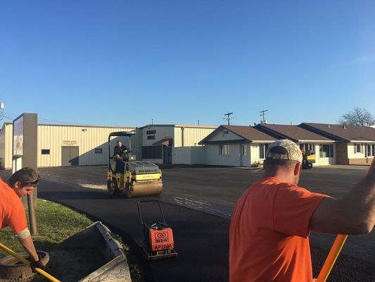 Workers paving asphalt with a roller and hand tools in front of a building on a sunny day.