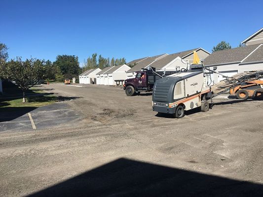 Parking lot with roadwork: truck, sweeper, and machinery in front of apartments on a sunny day.