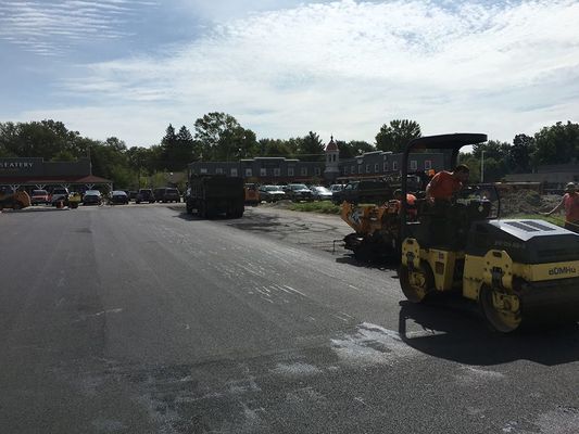 Construction workers paving a road; a steamroller and truck are on a fresh asphalt surface in a sunny setting.