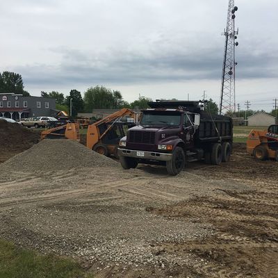 Construction site: dump truck unloading gravel, orange loaders, cloudy sky.