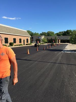 Freshly paved parking lot with orange cones, person in orange shirt. Buildings in background.