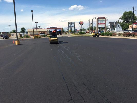 Asphalt paving in progress: a worker operating a roller, with businesses in the background under a blue sky.