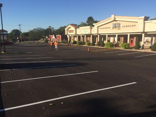 People riding bicycles in an empty parking lot in front of a strip mall, with businesses including a jewelry store.