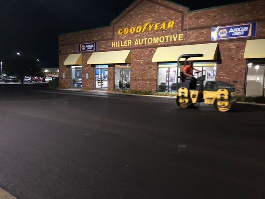 Asphalt roller smoothing new pavement in front of a Goodyear Hiller Automotive at night.