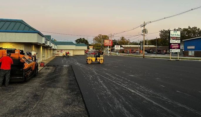 Asphalt paving a commercial parking lot with workers and machinery at dusk.
