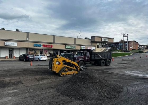A skid steer loading gravel into a dump truck in a shopping center parking lot.