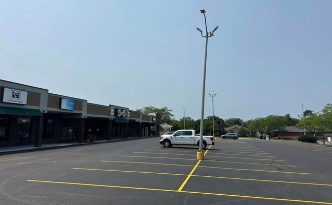 A parking lot with yellow lines and a white pickup truck, fronting a strip of stores under a blue sky.