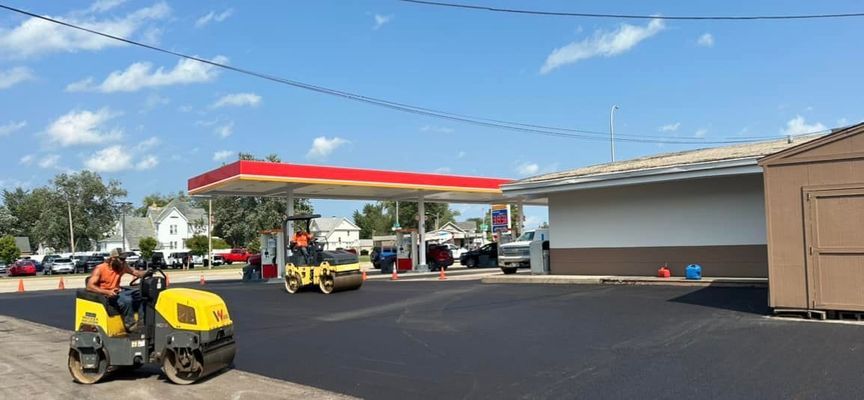 Asphalt paving at a gas station, with two yellow rollers working. Bright blue sky.