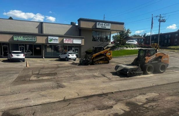 Two skid steer loaders working on a damaged asphalt parking lot in front of a strip mall.