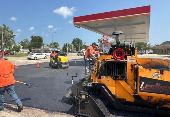 Asphalt paving at a gas station, workers in orange vests, yellow machinery, blue sky, and cars in the background.