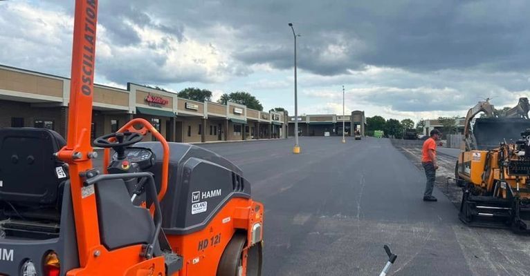 Asphalt paving a parking lot near a strip mall; construction workers operating machinery on a cloudy day.