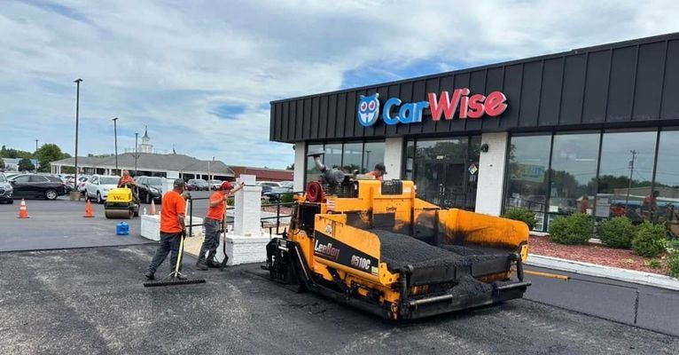 Asphalt paving at CarWise dealership. Workers in orange vests and a yellow paving machine.