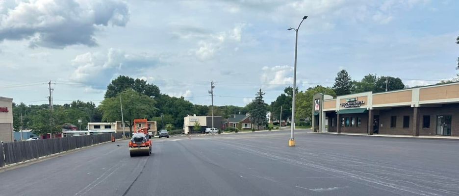 A road roller compacts fresh asphalt on a street lined with buildings under a cloudy sky.