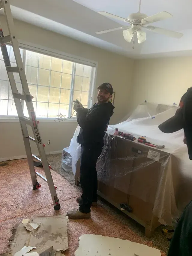 A man is standing in a room with a ladder and a ceiling fan.