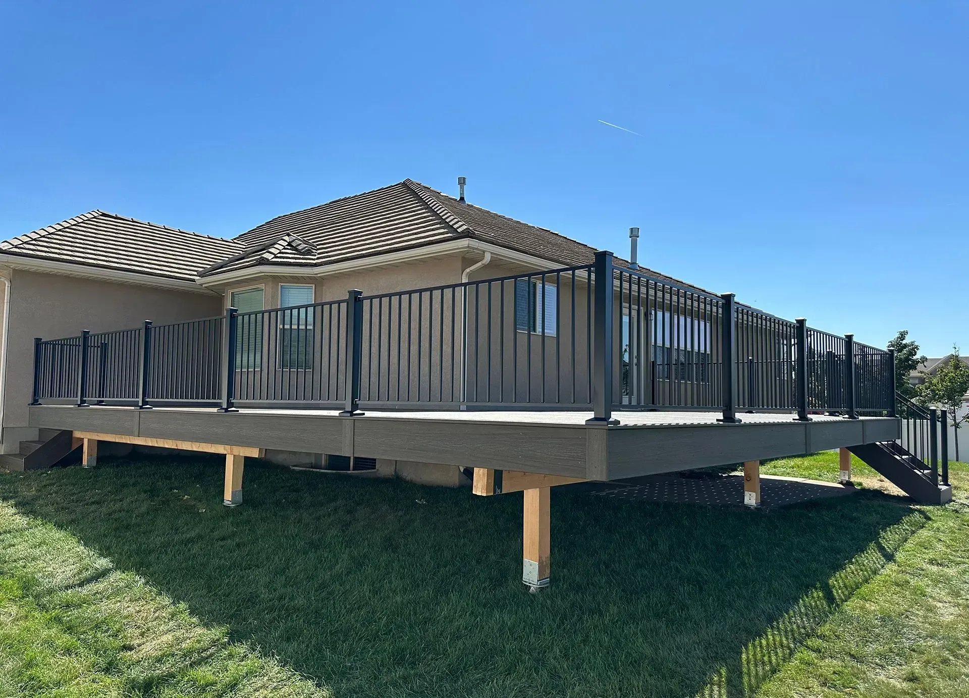 Elevated deck with black railings attached to a house with tan walls and a brown roof; set on a grassy lawn.